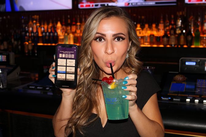 Woman in a dim bar sipping a blue-green cocktail with cherries while holding a smartphone showing an app, with bottles and slot machines in the background.