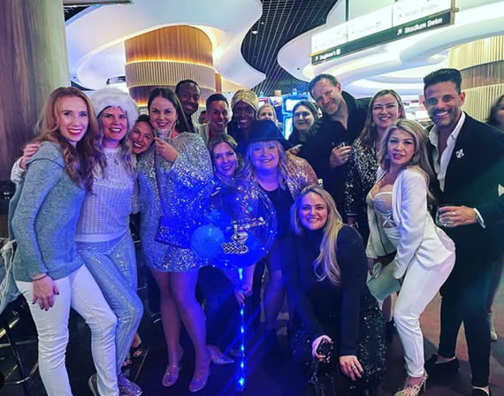 Group of friends in sparkly outfits posing with a glowing blue LED balloon and drinks inside a modern casino-style nightlife venue with curved ceiling lights.
