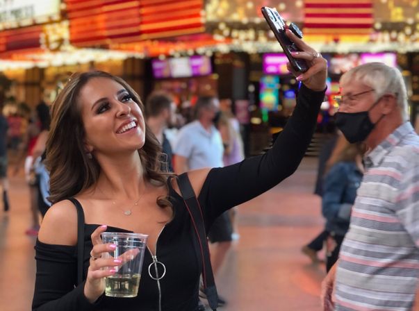 Young woman in a black off-shoulder top smiling and taking a selfie with her phone while holding a plastic cup, under bright marquee lights in a busy nighttime entertainment district.