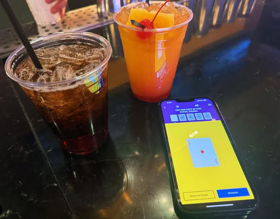 Two plastic bar drinks — a dark soda with ice and a bright orange cocktail garnished with an orange slice and cherry — next to a smartphone showing a digital card game on a glossy bar counter, nightlife vibe.