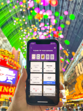 Hand holding a smartphone showing a tip/check-in app screen beneath a colorful LED canopy and neon signs in a lively Las Vegas-style entertainment district, with blurred green, pink and orange bokeh lights.