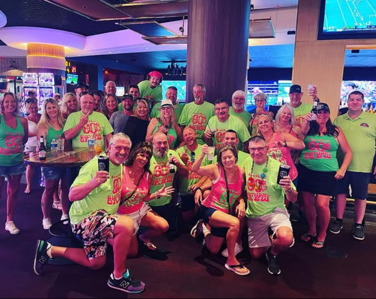 Large group of adults in bright neon green and pink '60th birthday' shirts posing and cheering with drinks inside a casino bar, slot machines and TV screens visible in the background.
