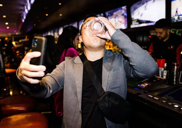 Patron taking a selfie while chugging a drink at a busy sports bar with multiple TV screens and a lit bar counter