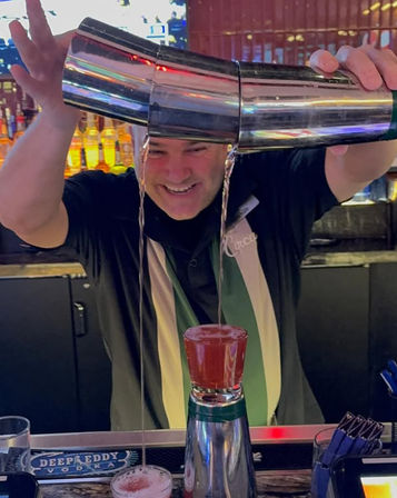 Smiling bartender at a cocktail bar pouring twin streams from stacked metal shakers into a layered shot glass on the counter, colorful backlit liquor bottles in the background.