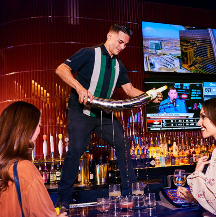 Bartender standing on the bar performing a dramatic long pour from a curved metal pitcher into multiple cocktail glasses as two patrons smile, with backlit liquor shelves and TV screens in the lively upscale bar scene.