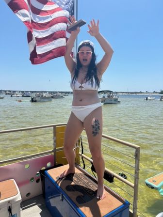 Woman in white bikini and backward cap standing on a pontoon platform under a waving American flag, cheering with arms raised while boats dot a sunny bay
