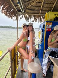 Two smiling women in bikinis and matching caps hugging on a tiki-roofed pontoon boat over green bay water, with a life ring, yellow equipment and tree-lined shore visible — summer boat outing.
