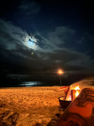 Cozy moonlit beach bonfire on a sandy shore, ocean reflecting moonlight under a cloudy night sky with a lone lamp