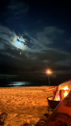 Cozy moonlit beach bonfire on a sandy shore, ocean reflecting moonlight under a cloudy night sky with a lone lamp