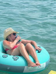 Woman in a red bikini and straw sun hat wearing sunglasses, sipping a canned drink while lounging on a turquoise inflatable float tube on calm teal water