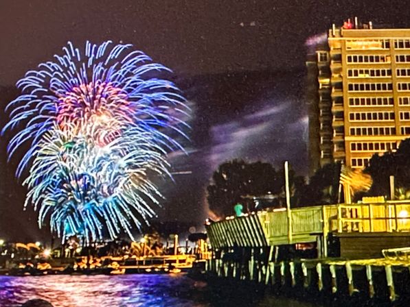 Vibrant fireworks burst over a waterfront pier at night, colorful blue and purple reflections on the water beside a lit high-rise and wooden boardwalk.
