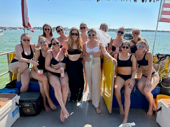 Smiling group of friends in swimsuits on a sunny boat bachelorette party in a coastal bay, bride wearing a veil with American flags and shoreline in the background.