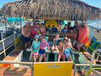 Smiling group of adults and children in swimsuits on a thatched‑roof tiki pontoon boat docked at a sunny coastal marina with boats and blue water in the background.