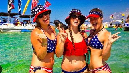 Three friends in American-flag bikinis and bucket hats laughing and posing in shallow turquoise beach water, with boats and colorful flags in the sunny background at a summer beach party