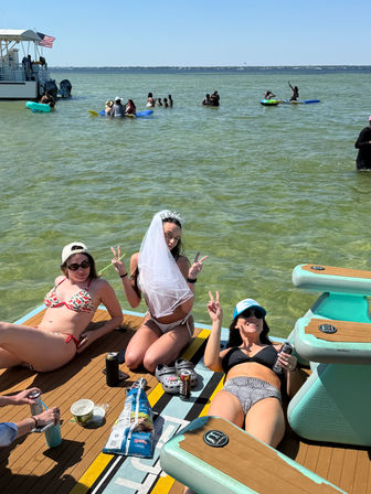 Three friends in bikinis relaxing on a floating pontoon in shallow green coastal water on a sunny day — bride-to-be with veil flashing peace signs, drinks and snacks nearby, paddleboarders and a small boat with an American flag in the background.