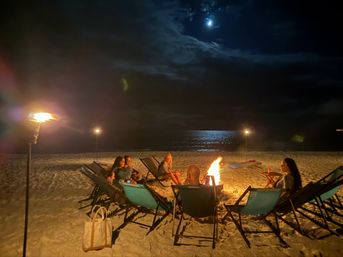 Moonlit beach bonfire — a small group relaxes in lounge chairs around a crackling fire, tiki torches lighting the sandy shore and moonlight shimmering on the ocean.