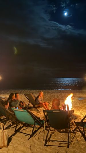 Moonlit beach bonfire — a small group relaxes in lounge chairs around a crackling fire, tiki torches lighting the sandy shore and moonlight shimmering on the ocean.