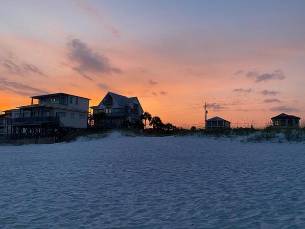 Silhouetted beach houses and palm trees on a sand dune at sunset, pastel pink-orange sky above rippled white sand shoreline