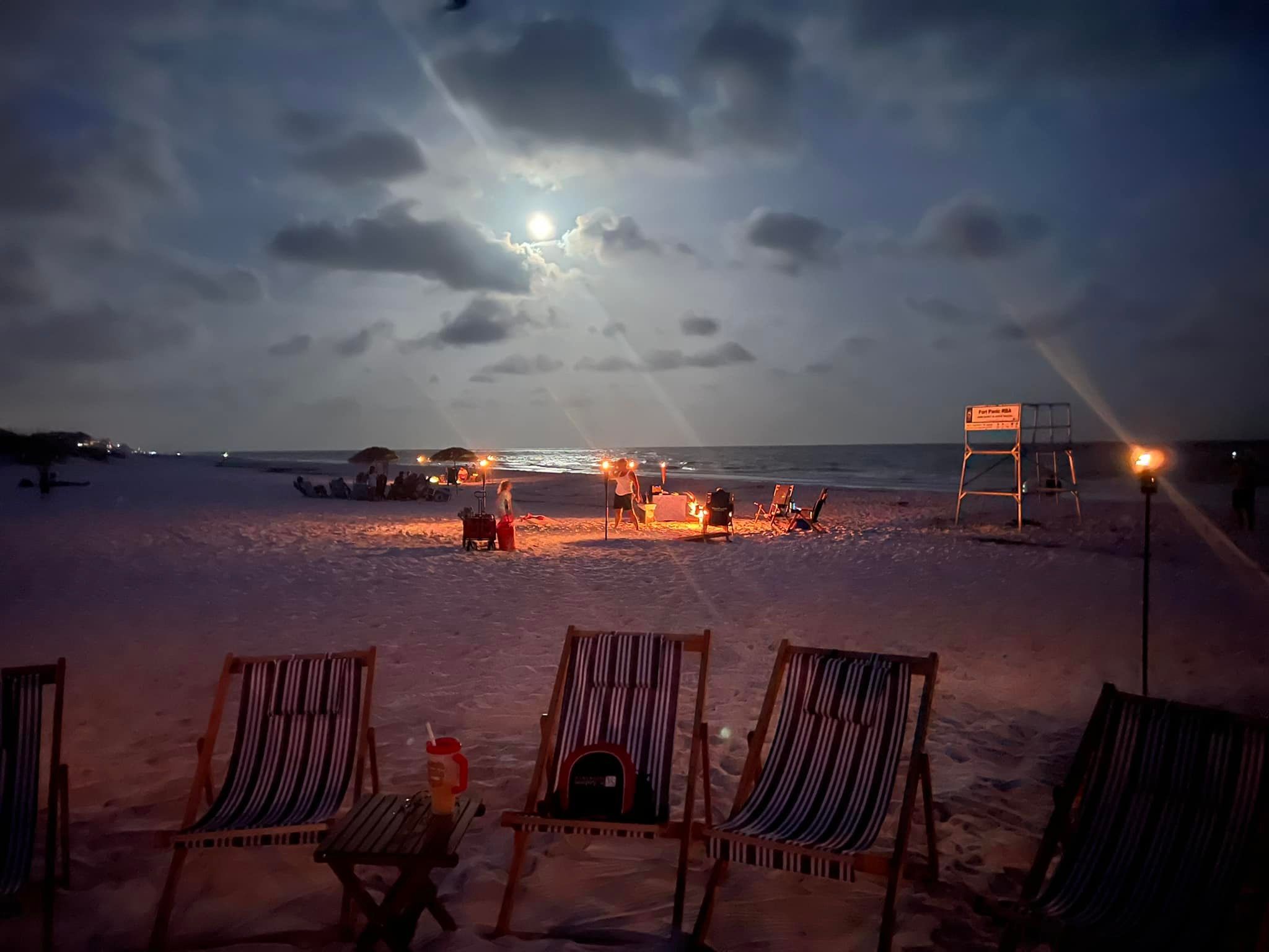 Moonlit beach at night with striped lounge chairs in the foreground, a small bonfire and groups gathered along the shoreline, tiki torches and a lifeguard stand under a cloudy sky reflecting on the ocean.