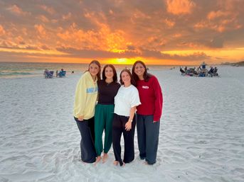 Four friends smiling and barefoot on a white‑sand beach at a vibrant orange sunset over the ocean, with beachgoers and chairs in the background.