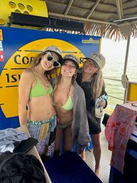Three smiling friends in green bikinis and matching caps posing under a thatched-roof on a tropical island boat with yellow signage and ocean in the background, beach vacation vibe.