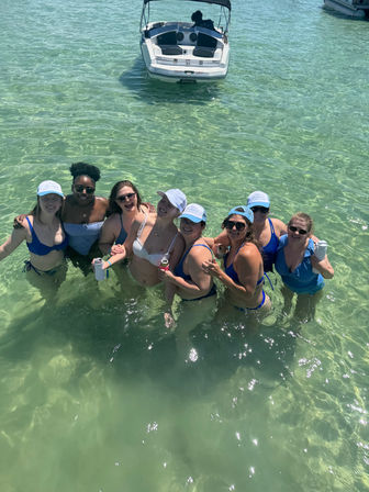 Eight women in bikinis and sun hats standing in shallow turquoise coastal water, holding drinks and smiling with an anchored motorboat in the background on a sunny day