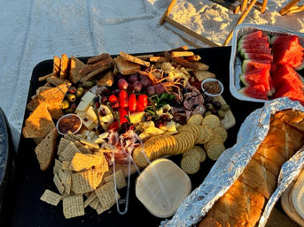 Colorful beach picnic charcuterie board on a black table over white sand: crackers, artisan bread, cheeses, cured meats, grapes, cherry tomatoes, olives, dips, and a tray of sliced watermelon at sunset.