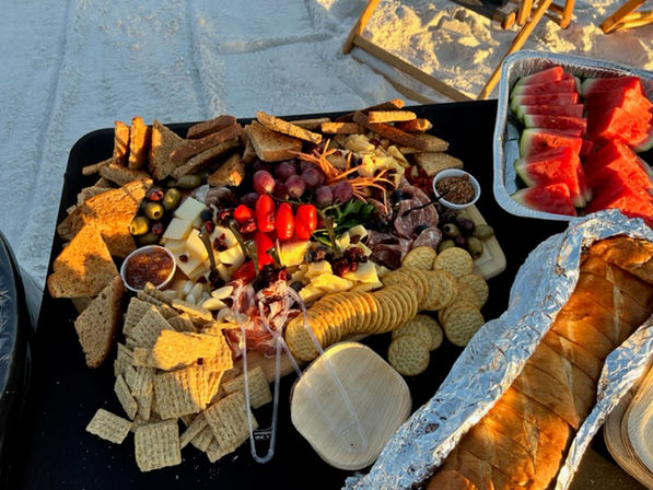 Colorful beach picnic charcuterie board on a black table over white sand: crackers, artisan bread, cheeses, cured meats, grapes, cherry tomatoes, olives, dips, and a tray of sliced watermelon at sunset.