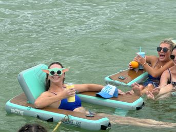 Three women in blue swimsuits relaxing on turquoise inflatable loungers in calm green water, laughing and sipping drinks; one wears playful round sunglasses with ear-like attachments.