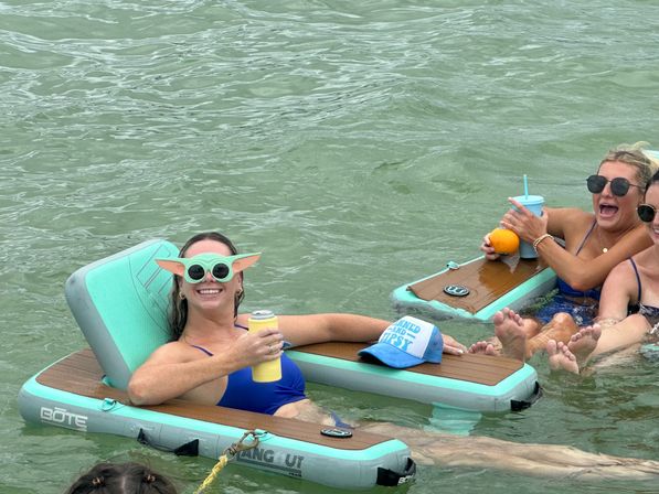 Group of friends relaxing on turquoise inflatable floating loungers in green water, laughing and holding cold drinks — one wearing playful green alien‑ear sunglasses.