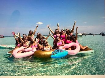 Group of women in pink swimsuits and heart-shaped sunglasses cheering on a large paddleboard with pink flamingo and gold donut inflatables in clear turquoise water with boats and a sunny sky