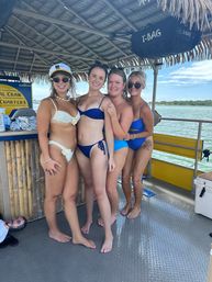 Four friends in colorful bikinis smiling and posing under a thatched-roof tiki on a sunny coastal boat with calm bay water in the background.