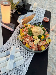 Beach picnic seafood boil on a sandy beach: large pan piled with shrimp, corn on the cob, red potatoes, a whole cauliflower, lemon wedges and parsley, with a foil-wrapped loaf, dipping sauce, stacked napkins and a glowing lantern on a black table.