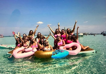 Group of women in pink swimsuits and heart-shaped sunglasses on a floating platform with pastel inflatable rings in clear turquoise shallow water under a bright blue sky — lively beach party scene.