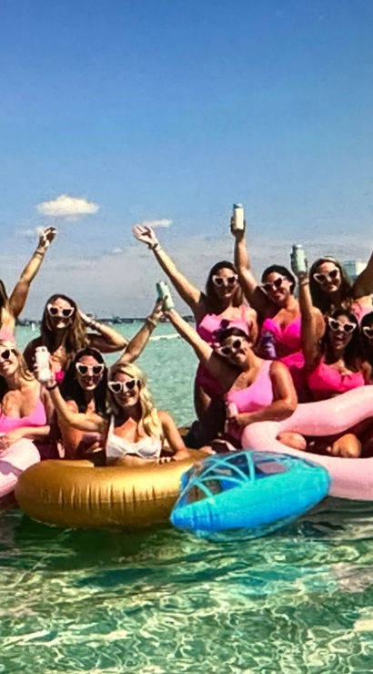 Group of women in pink swimsuits and heart-shaped sunglasses on a floating platform with pastel inflatable rings in clear turquoise shallow water under a bright blue sky — lively beach party scene.