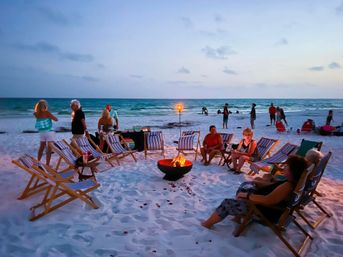 Dusk beach scene with people relaxing around a small bonfire on white sand, striped deck chairs in a circle, a glowing tiki torch, and calm ocean waves with others strolling along the shoreline.