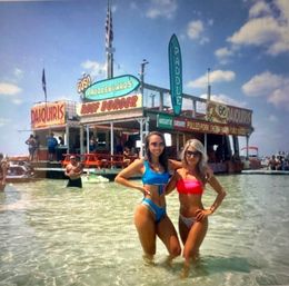 Two friends in colorful bikinis posing waist-deep in clear shallow beach water in front of a lively floating beach bar with paddleboard and daiquiri signs on a sunny day.