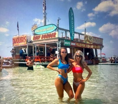 Two women in colorful bikinis standing waist‑deep in clear shallow beach water in front of a lively floating beach bar and rental shack with signs for daiquiris, burgers and paddleboards on a sunny day.