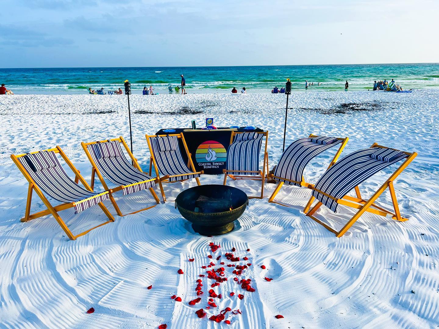 White-sand beach with six striped wooden lounge chairs in a semicircle around a metal fire pit and tiki torches, red rose petals scattered toward the pit, turquoise ocean and people along the shoreline.