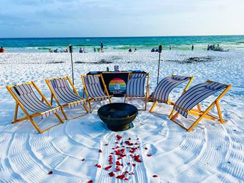 White-sand beach with six striped wooden lounge chairs in a semicircle around a metal fire pit and tiki torches, red rose petals scattered toward the pit, turquoise ocean and people along the shoreline.