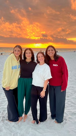Four friends smiling and barefoot on a white‑sand beach at a vibrant orange sunset over the ocean, with beachgoers and chairs in the background.