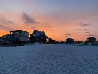 Silhouetted beach houses and palm trees on a sand dune at sunset, pastel pink-orange sky above rippled white sand shoreline