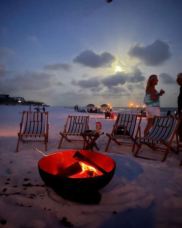 Moonlit beach bonfire at dusk with a glowing fire pit in the sand, striped lounge chairs, tiki torches and silhouetted people gathered along the shoreline beneath a cloud-covered moon.
