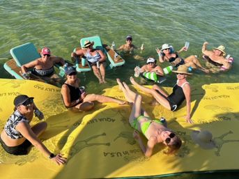 Group of women in swimsuits and sun hats relaxing on a large yellow floating mat and nearby lounge floats in shallow green water, holding drinks and enjoying a sunny summer lake/beach float party.