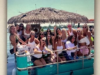 Group of women enjoying a sunny lake cruise on a turquoise pontoon boat with a thatched tiki canopy, wearing sunglasses and holding drinks.