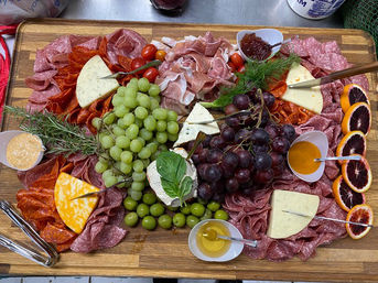 Vibrant wooden charcuterie board piled with assorted cured meats, mixed cheeses, green and red grapes, green olives, blood orange slices, fresh herbs, and small bowls of honey and jam.