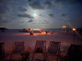 Moonlit beach at night with striped lounge chairs in the foreground, a small bonfire and groups gathered along the shoreline, tiki torches and a lifeguard stand under a cloudy sky reflecting on the ocean.