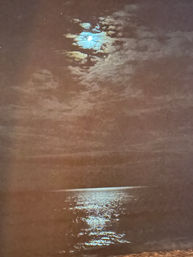 Moon peeking through clouds above a calm coastal night sea, casting a shimmering silver reflection across the water.