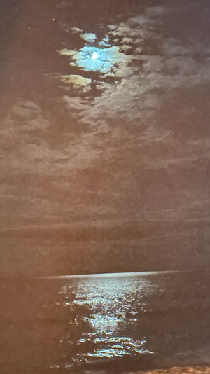 Moon peeking through clouds above a calm coastal night sea, casting a shimmering silver reflection across the water.