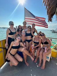 Group of friends in black swimsuits posing on a sunny boat deck during a summer boat party, smiling and holding tumblers with a large American flag waving behind them over blue lake waters.